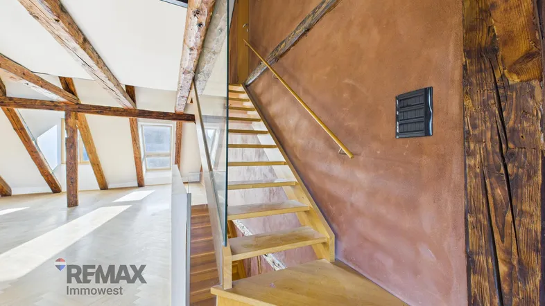 Rustikale Holztreppe mit Glasgeländer führt zu einem hellen Dachgeschoss mit sichtbaren Holzbalken.