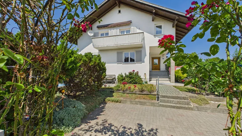 Weißes Einfamilienhaus mit Satteldach, kleinem Vorgarten und gepflasterter Auffahrt, umgeben von Rosenbüschen.
