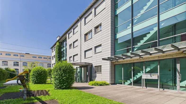 Modernes Bürogebäude mit Glasfassade und gepflegter Grünfläche im Eingangsbereich unter blauem Himmel.
