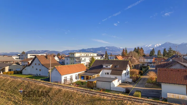 Luftaufnahme eines Einfamilienhauses mit Garten, Bahngleisen und Bergpanorama unter klarem Himmel.