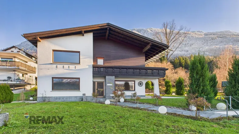 Haus mit weißer Fassade, Holzelementen und gepflegtem Vorgarten vor einer Bergkulisse.