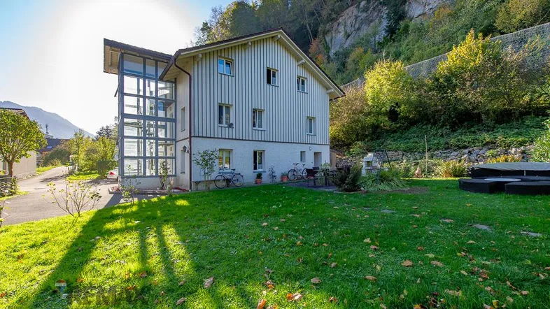 Großzügiger Gartenbereich vor dem Haus mit Rasenfläche und Sitzgelegenheit, umgeben von grüner Natur.
