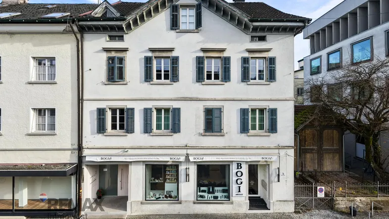 Klassisches Gebäude mit weißer Fassade, grünen Fensterläden und Ladenlokal im Erdgeschoss.