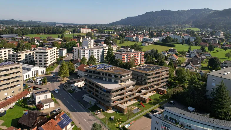 Luftaufnahme einer modernen Wohnanlage mit Flachdächern und Photovoltaikanlagen, umgeben von grüner Landschaft und Bergen.