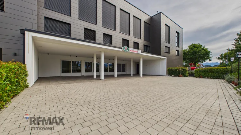 Modernes Gebäude mit überdachtem Eingangsbereich und gepflastertem Vorplatz, umgeben von Grünflächen.