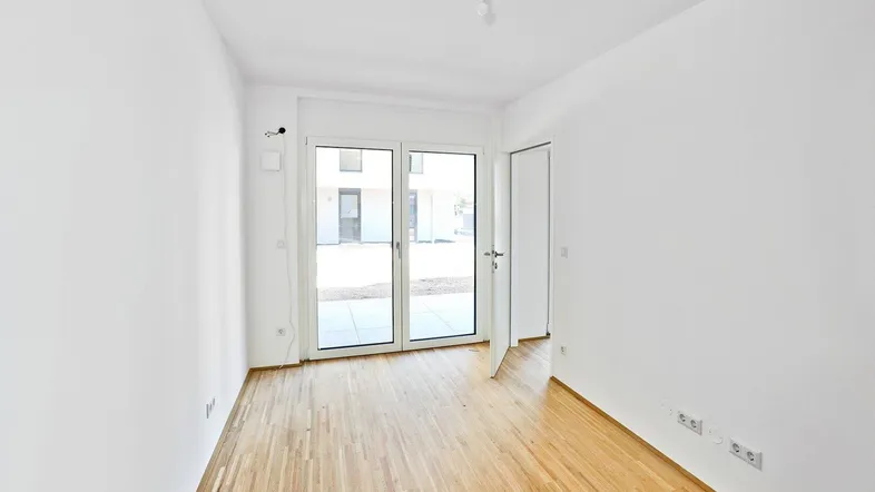 Heller, leerer Raum mit Parkettboden und Zugang zu einem Außenbereich durch große Glastüren.
