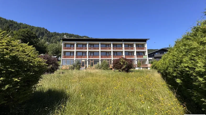 Gebäudeansicht mit mehreren Balkonen, umgeben von einer grünen Wiese und Bergen im Hintergrund.