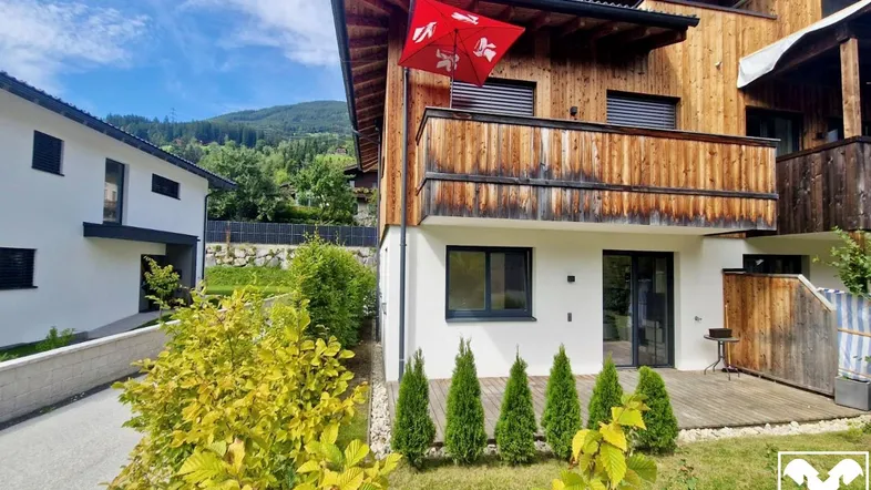 Modernes Gebäude mit Holzfassade und weißem Putz, umgeben von Grün und Bergblick.