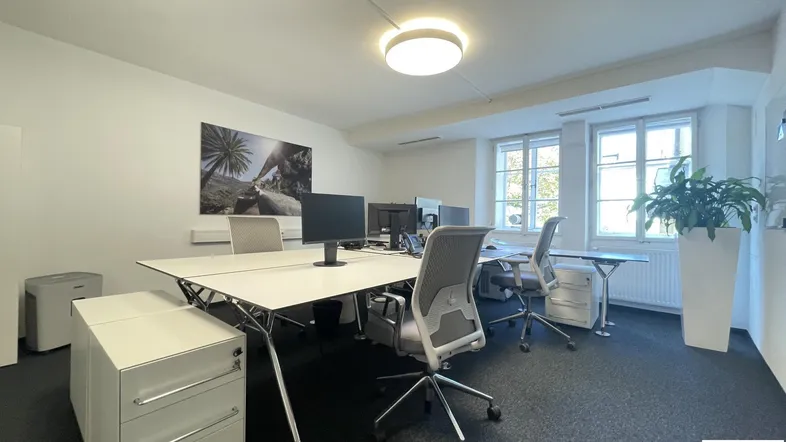 Helles und funktionales Büro mit mehreren Arbeitsplätzen, großen Fenstern und Teppichboden.