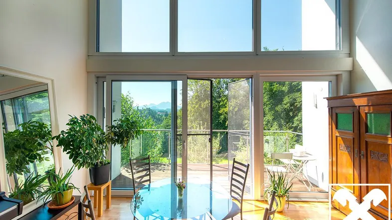 Heller Wohn- und Essbereich mit großen Fenstern, Holzboden und Zugang zu einem Balkon mit Bergblick.