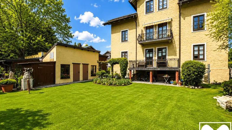 Gepflegter Garten mit großer Rasenfläche vor einem charmanten gelben Gebäude mit Balkon und Terrasse.