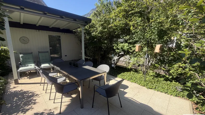 Überdachte Terrasse mit Esstisch und Stühlen, umgeben von viel Grün und Bergblick.