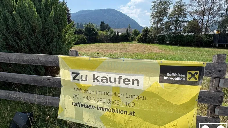 Gelbes 'Zu kaufen'-Banner an einem Holzzaun vor einem leeren Grundstück mit Bergblick.