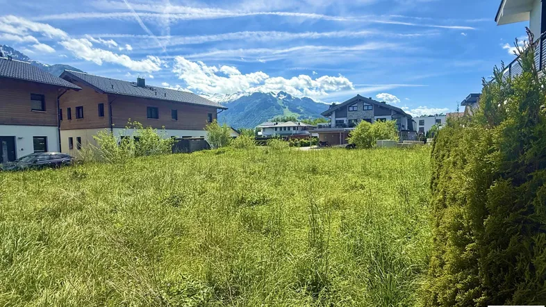 Blick über ein grünes Grundstück mit Wohnhäusern und einer beeindruckenden Bergkulisse unter blauem Himmel.