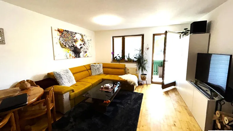 Helles Wohnzimmer mit gelbem Ecksofa, Holzboden und Zugang zum Balkon.
