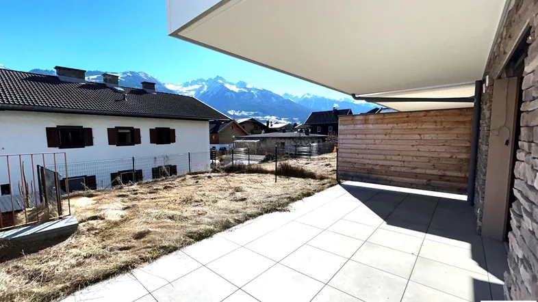 Große geflieste Terrasse mit Bergblick und angrenzender Grünfläche unter blauem Himmel.