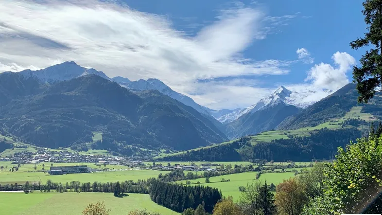 Panoramablick auf eine weite grüne Tallandschaft mit majestätischen Bergen und einem Dorf im Hintergrund.