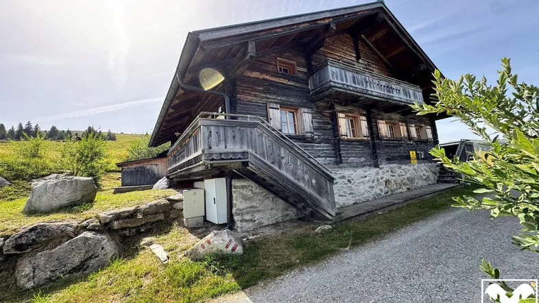 Rustikales Holzchalet mit Steinfundament und Balkon in ländlicher Umgebung, umgeben von grünen Hügeln.