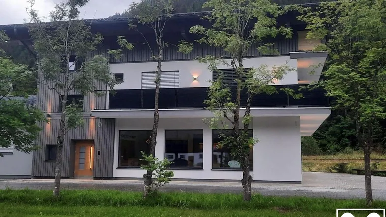 Modernes Gebäude mit Holz- und Putzfassade, Balkonen und großen Fenstern in bergiger Umgebung.