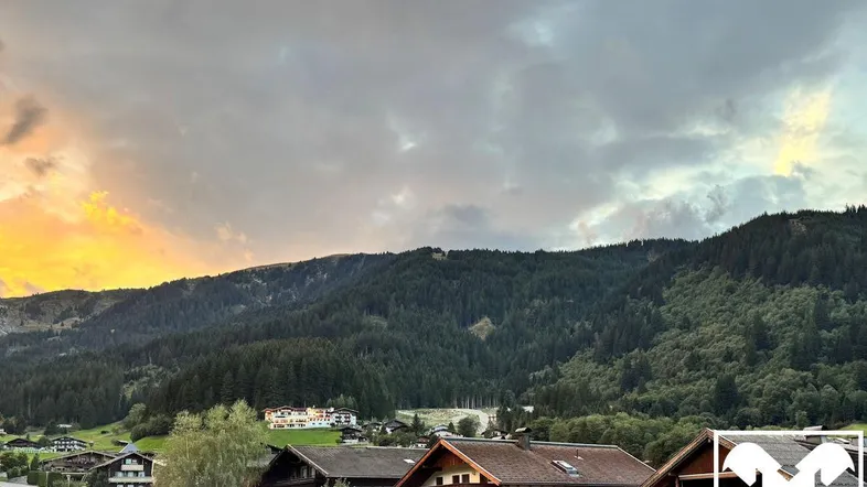 Panoramablick auf ein malerisches Dorf mit traditionellen Häusern, eingebettet in eine bewaldete Berglandschaft unter einem dramatischen Abendhimmel.