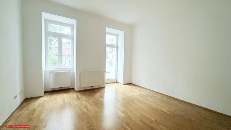 Helles Zimmer mit zwei Fenstern, weiß gestrichenen Wänden und Parkettboden.