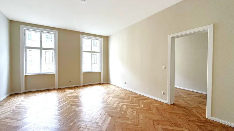 Helles Wohnzimmer mit Fischgrätparkett und zwei großen Fenstern, die viel Tageslicht hereinlassen.
