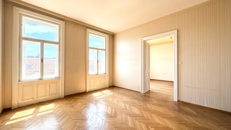 Helles Zimmer mit Fischgrätparkett, hohen Decken und großen Fenstern, die viel Tageslicht hereinlassen.