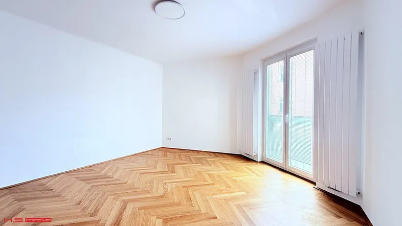 Helles Zimmer mit Fischgrätparkettboden, weißen Wänden und Zugang zu einem französischen Balkon.