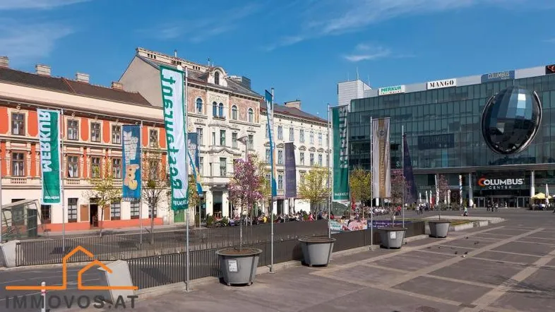 Blick auf eine belebte Straße mit historischen Gebäuden und einem modernen Einkaufszentrum unter blauem Himmel.