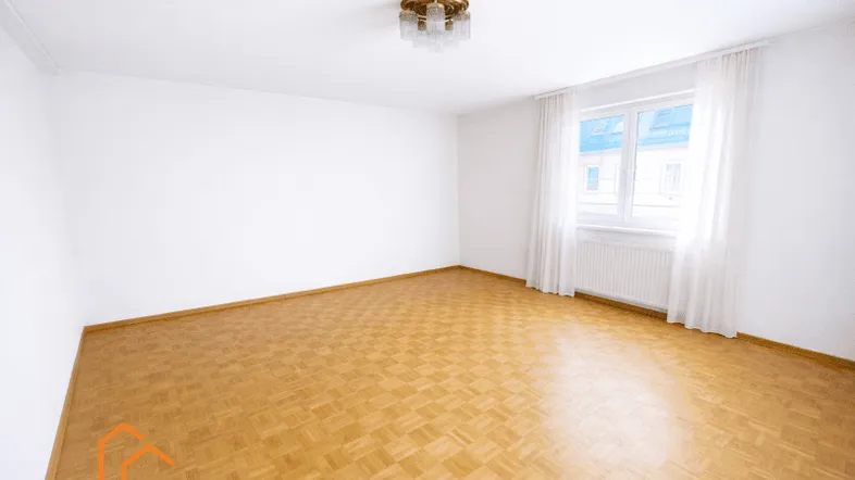 Helles, leeres Zimmer mit Parkettboden und einem Fenster, das viel Tageslicht hereinlässt.