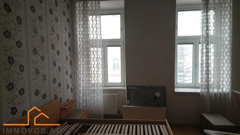 Schlafzimmer mit gemusterter Tapete, Holzlattenrost und zwei Fenstern für natürliches Licht.