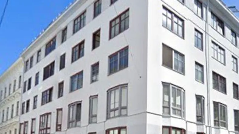 Eckgebäude mit weißer Fassade und vielen Fenstern an einer städtischen Straße unter blauem Himmel.