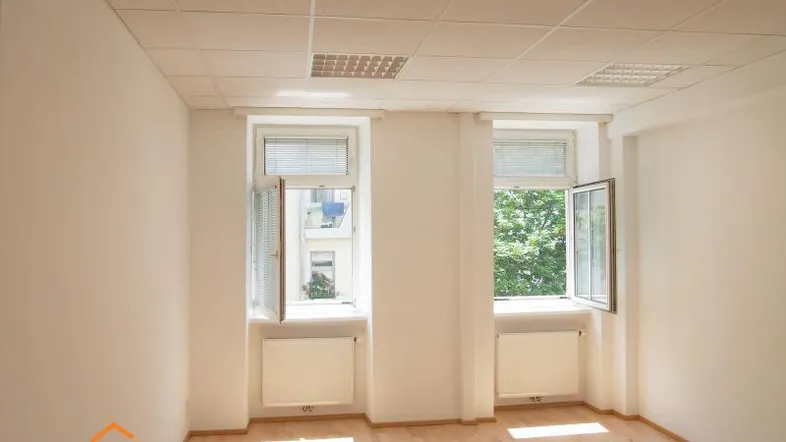 Helles, leeres Zimmer mit zwei Fenstern, Holzfußboden und Rasterdecke, ideal für Büro oder Wohnzwecke.