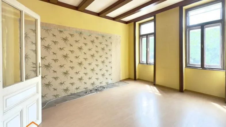 Helles Zimmer mit gelben Wänden, sichtbaren Deckenbalken und Laminatboden, bereit zur Renovierung.