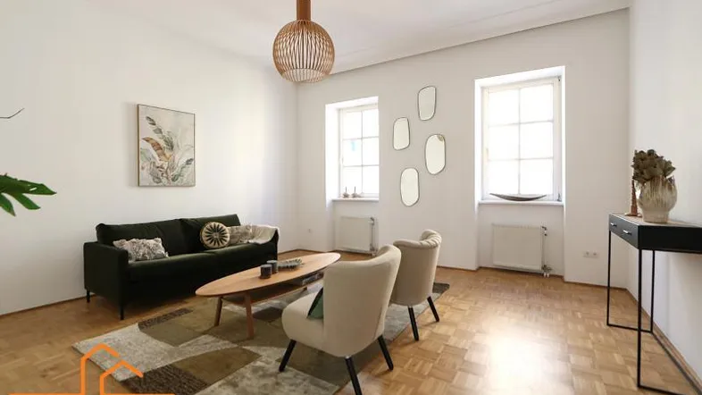Helles Wohnzimmer mit grünem Sofa, zwei Sesseln und Fischgrätparkettboden, durch zwei Fenster gut belichtet.