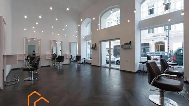 Geräumiger, moderner Friseursalon mit hohen Decken, großen Fenstern und dunklem Fischgrätparkett.