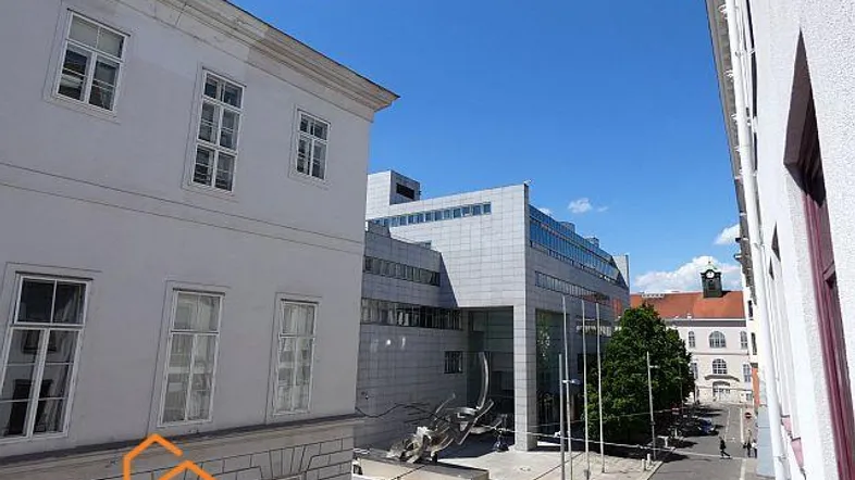 Blick auf die Straße mit verschiedenen Gebäuden und einem modernen Bau im Hintergrund unter blauem Himmel.