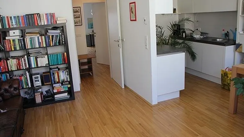 Heller Wohnbereich mit Holzfußboden, Bücherregal und offenem Übergang zur Küche.