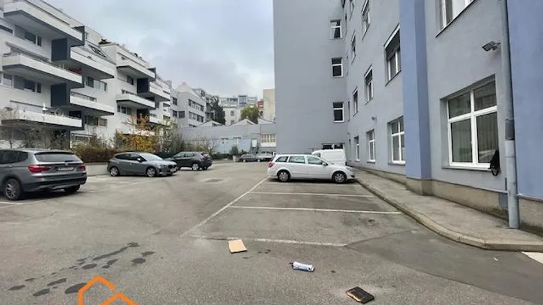 Außenansicht eines Parkplatzes mit mehreren geparkten Autos und umliegenden Gebäuden unter bewölktem Himmel.