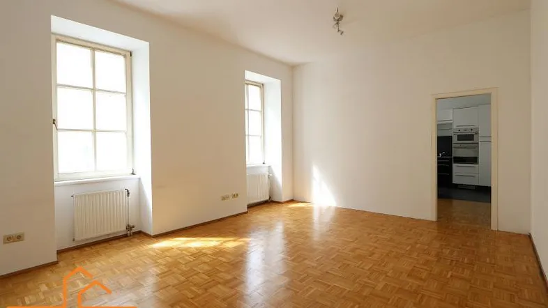 Helles, leeres Zimmer mit Fischgrätparkettboden und zwei hohen Fenstern, ideal als Wohnzimmer.