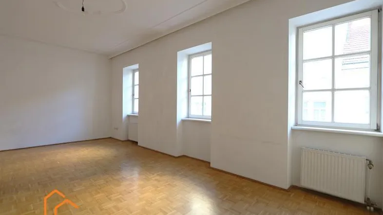 Helles Zimmer mit Fischgrätparkett, hohen Fenstern und dekorativer Stuckdecke, ideal als Wohnzimmer.