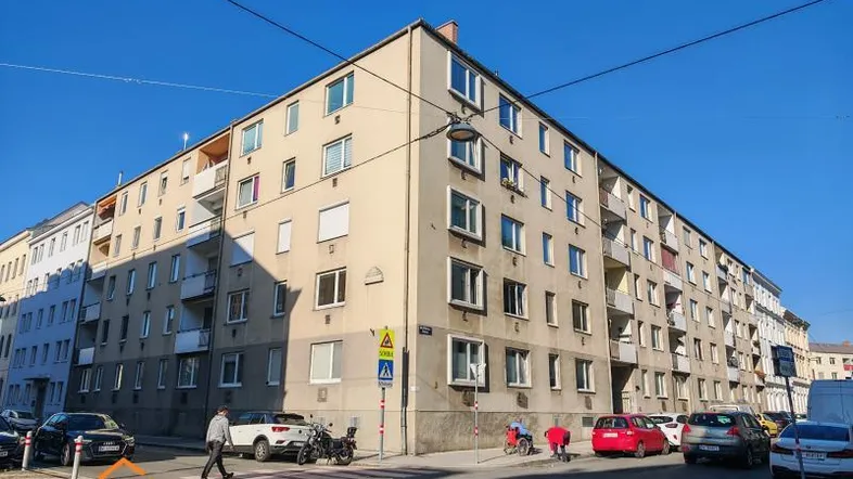 Mehrstöckiges Wohngebäude an einer Straßenecke mit parkenden Autos und Fußgängern.