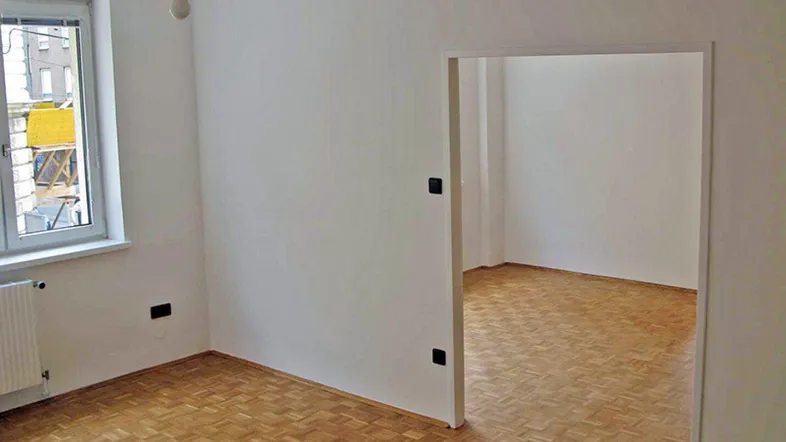 Helles, leeres Zimmer mit Parkettboden und Fenster, das einen Blick auf die Umgebung bietet.