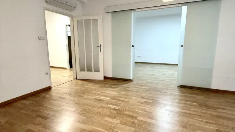 Heller Raum mit Holzparkettboden, weißen Wänden und einer Glasschiebetür zu einem weiteren Zimmer.