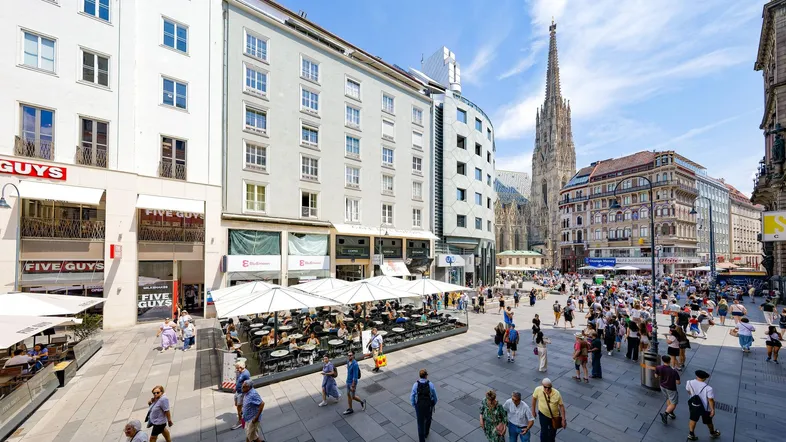 Belebter Stadtplatz mit historischen Gebäuden, Cafés und dem Stephansdom im Hintergrund.