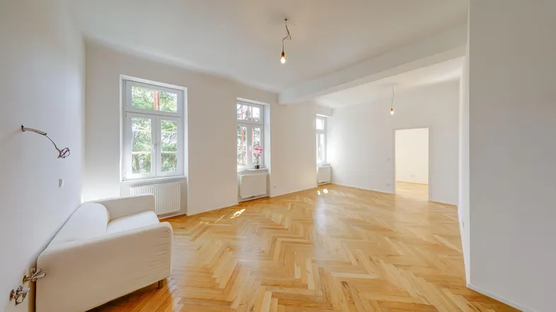 Helles Zimmer mit Fischgrätparkett, weißen Wänden und großen Fenstern, ideal als Wohnbereich.
