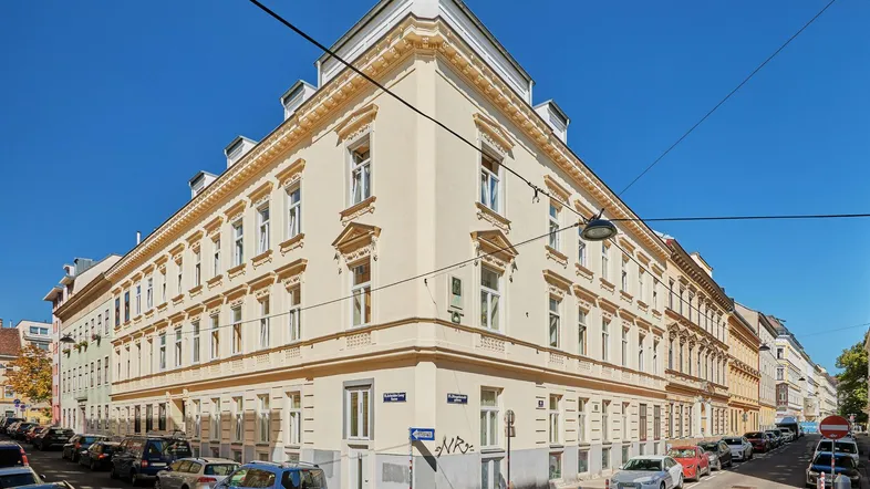 Eckgebäude mit klassischer, gut erhaltener Fassade und vielen Fenstern an einer belebten Stadtstraße.