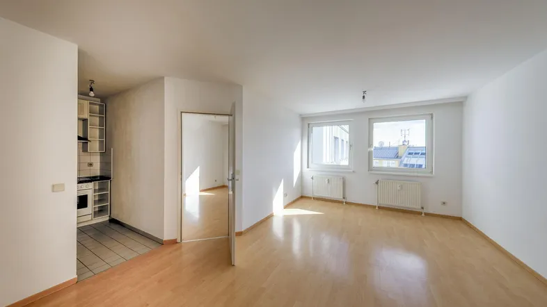 Helles Wohnzimmer mit Parkettboden, weißen Wänden und Blick nach draußen, angrenzend an eine Küchennische.