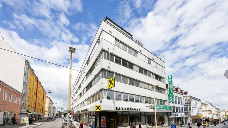 Gebäudeansicht an einer belebten Straße mit Geschäften und Passanten unter blauem Himmel.