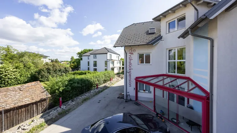 Vorderansicht eines Gebäudes mit rotem Wintergarten und geparktem Auto unter blauem Himmel.